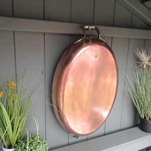 May include: A large, oval-shaped copper pan with a polished, reflective surface and a single curved handle at the top. The pan is constructed from smooth, warm-toned copper metal and features a simple, functional design for culinary use.