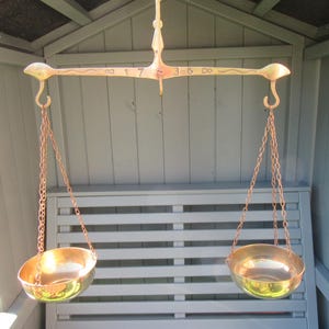 May include: Vintage brass balance scale featuring two circular bowls hanging from chains attached to a curved arm. The arm is inscribed with the numbers "17" and "36". The scale is set against a grey wooden backdrop.