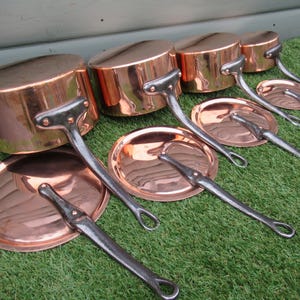 May include: A set of copper cooking pots, pans, and lids. The copper has a polished finish, and the handles are made of a darker metal. The cookware is displayed on a green surface.