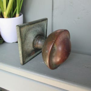 May include: A vintage door knob featuring a rounded, weathered copper-toned handle attached to a square, oxidized metal backplate. The hardware displays a distinct patina with visible signs of age, including dark spots and a dull, matte finish.