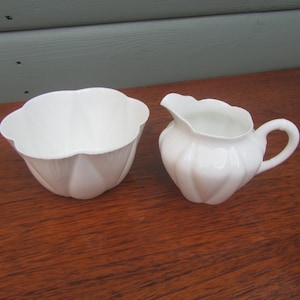 Op de afbeelding: Een witte porseleinen melk- en suikerpotset. De melkkan heeft een handvat en een tuit, terwijl de suikerpot een geschulpte rand heeft. Beide stukken hebben een geribbeld ontwerp en staan op een houten oppervlak.