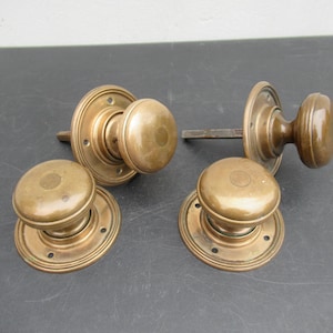 May include: Four brass door knobs with circular backplates. Two of the knobs feature attached square spindles extending from the back, while the other two are standalone knobs with circular mounting plates featuring small screw holes.
