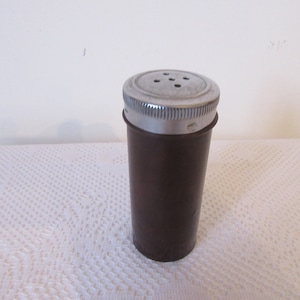 Può includere: Uno shaker di plastica marrone con un coperchio argentato e fori per la distribuzione. Lo shaker ha una forma rotonda e liscia.