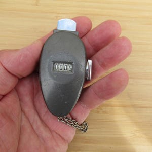 Può includere: Un contatore meccanico manuale con un involucro metallico ovale di colore grigio scuro. Il dispositivo presenta una piccola finestra di visualizzazione che mostra il numero 0005, un pulsante argentato sulla parte superiore, una manopola di reset laterale e una corta catenella metallica.