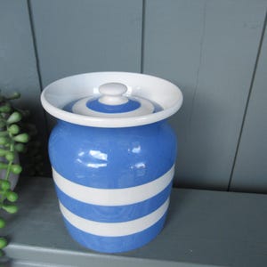 May include: A blue and white striped ceramic jar with a lid. The jar has a round shape with horizontal white stripes against a blue background. The lid is white with a blue and white circular design. The jar is sitting on a gray surface.