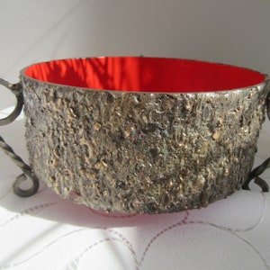 May include: A rustic-looking metal bowl with a red interior and twisted metal handles. The bowl has a textured, bark-like exterior.
