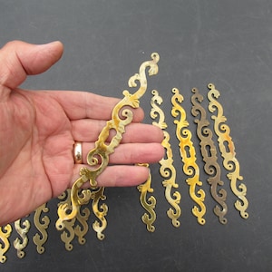 Könnte beinhalten: Eine Sammlung länglicher Schlüsselschilder aus Messing mit einem verzierten, geschwungenen Design und einer zentralen Schlüssellochöffnung. Jedes Metallschild hat eine polierte goldfarbene Oberfläche und geschwungene, dekorative Kanten.