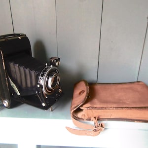 May include: A classic black folding camera with a silver lens and a brown leather case. The camera is open, showing the bellows and lens. The case is rectangular with a flap and strap closure.