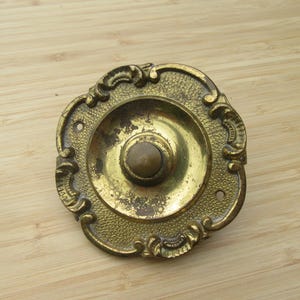 May include: Ornate brass door bell button with a central button and decorative scrollwork around the perimeter. The antique doorbell has a textured surface and a tarnished finish, suggesting age and use.