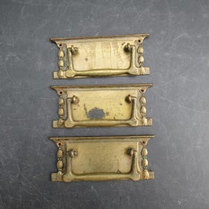 May include: Three antique brass drawer pulls with a rectangular design and curved handles. Each pull features decorative beaded accents on the sides. The brass has a weathered, aged appearance, with some areas showing wear and discoloration.