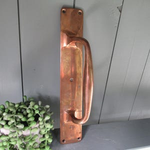 May include: A vintage copper door pull with a rectangular backplate and a curved handle. The door pull has a polished finish and is mounted on a gray wooden surface. The handle is a warm copper color.