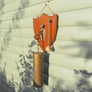 May include: A vintage wall-mounted barometer with a shield-shaped wooden backing. The barometer features brass and gold-toned metal accents, including a decorative hook, a pendulum, and a cylindrical weight. The wooden shield is a warm brown colour.