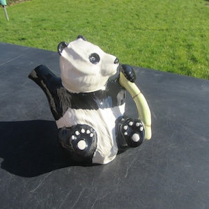 Puede incluir: Una tetera de cerámica negra y blanca con forma de panda que sostiene un trozo de bambú en la boca.