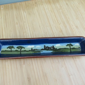 Puede incluir: Una bandeja rectangular larga de cerámica con un interior azul profundo y un borde exterior marrón. El interior presenta un paisaje pintado a mano que representa campos verdes, un cuerpo de agua, árboles dispersos y estructuras lejanas bajo el cielo.