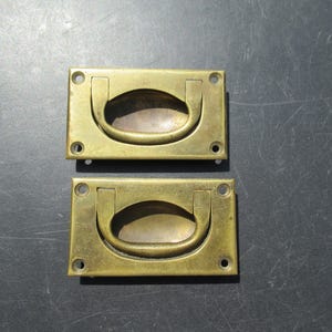 May include: Two antique brass recessed drawer pulls. Each rectangular pull has a curved handle and four screw holes. The brass has a warm, aged patina. The pulls are set against a dark background.