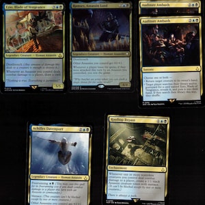 May include: Five Magic: The Gathering cards depicting assassins. The cards are "Ezio, Blade of Vengeance," "Ramses, Assassin Lord," "Auditore Ambush," "Achilles Davenport," and "Rooftop Bypass." Each card has a distinct design and text, with various game effects.