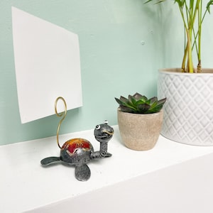 May include: A metal turtle photo holder with a gold wire loop. The turtle is painted with red, orange, and yellow accents. The turtle is holding a white card.