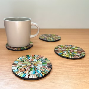 May include: A beige mug sits on a coaster with a mosaic pattern. Three more coasters with the same design are on a light wood surface. The coasters have a circular design with a black border and a vibrant mix of colours, perfect for a cuppa.