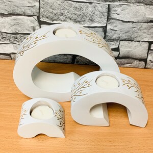 May include: Three white wooden candle holders with gold leaf designs. The holders are shaped like a crescent moon and have a small opening for a tea light candle.