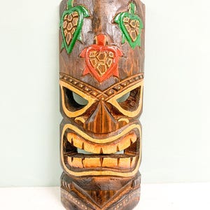 May include: A wooden tiki mask with a carved face and three green and red turtle designs. The mask is painted with brown, yellow, and green paint.