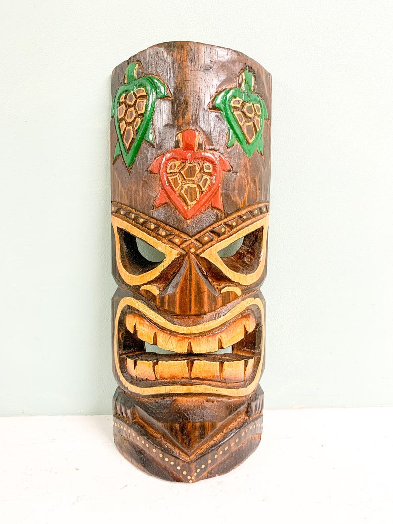 May include: A wooden tiki mask with a carved face and three green and red turtle designs. The mask is painted with brown, yellow, and green paint.