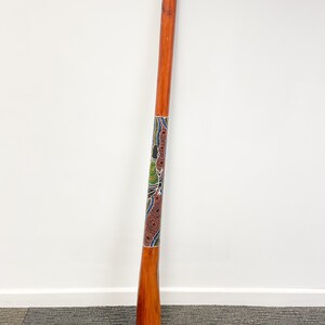May include: A wooden didgeridoo with a colorful Aboriginal art design. The didgeridoo is brown with a blue, green, red, and black painted design.