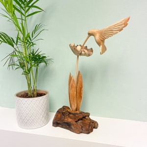 May include: A wooden hummingbird sculpture feeding from a carved flower, mounted on driftwood. The wings of the bird are outstretched. A potted plant is in the background.