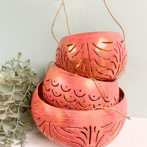 May include: Three pink hanging planters with intricate cut-out designs. The planters are stacked on top of each other and have a gold metallic finish.