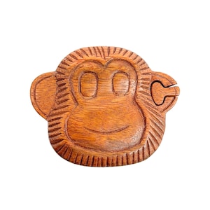 May include: A wooden monkey puzzle box with a carved face and a hinged ear. The box is made of light brown wood and has a natural finish.