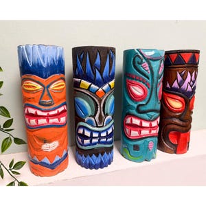 May include: Four brightly coloured, carved wooden tiki statues. Each statue has a unique design with vibrant colours including orange, blue, teal, and brown. The statues have detailed facial features and are displayed on a white shelf.
