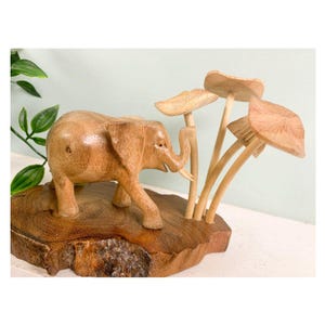 May include: A wooden sculpture featuring a carved elephant walking on a wooden base, accompanied by wooden mushroom figures. The elephant is light brown, with visible details. The mushrooms have a light brown color. The base has a natural wood grain.