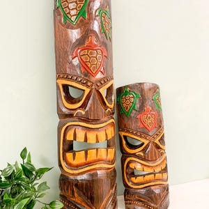 May include: Two wooden tiki masks with carved details. The larger mask has a brown, orange, and green color scheme with a carved face and three turtles. The smaller mask has a similar design with a brown, orange, and green color scheme.
