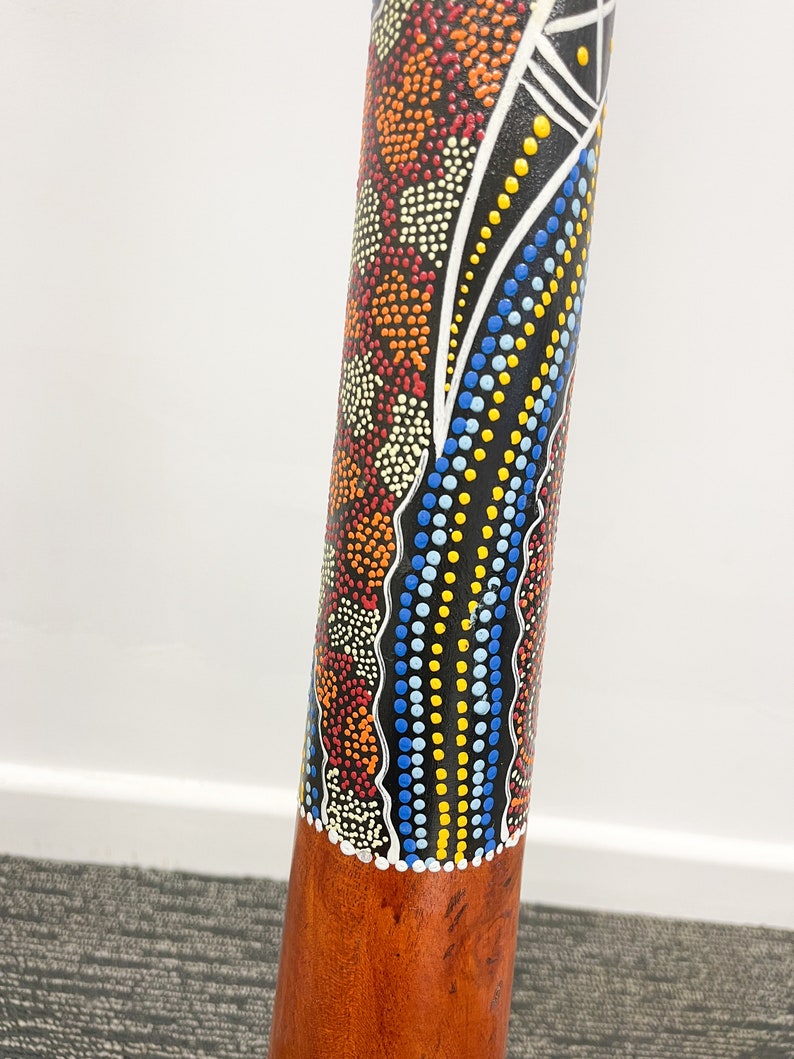 May include: A wooden didgeridoo with a black, white, blue, yellow, and orange painted design. The design features dots and wavy lines.