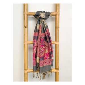May include: A gray scarf with a detailed pattern of elephants, pink and purple floral designs, and geometric borders. The scarf is draped over a bamboo ladder, showcasing its intricate details and fringed edges.