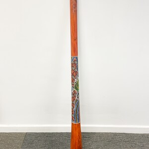 May include: A wooden didgeridoo with a colorful Aboriginal design painted on it. The didgeridoo is brown and has a natural wood finish. The design is a mix of red, blue, green, and yellow.