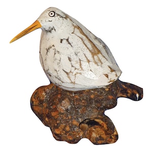 May include: A hand-carved wooden bird sculpture with a white and brown distressed finish. The bird has a long yellow beak and is perched on a textured, brown wooden base. The sculpture is a decorative item.