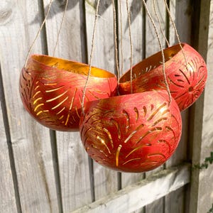 May include: Three hanging planters with a red exterior and gold interior. Each planter has a leaf-like cut-out design and is suspended by twine. The planters are set against a wooden fence.