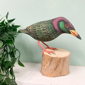 May include: Hand-painted wooden bird sculpture with green, purple and black feathers. The bird is perched on a small wooden stump.