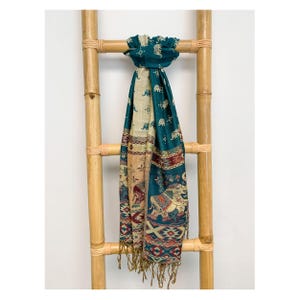 May include: A teal scarf with an elephant pattern and fringe hangs on a bamboo ladder. The scarf features a repeating elephant design, geometric patterns, and a beige and teal color scheme. The scarf is a decorative accessory.
