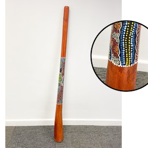 May include: A wooden didgeridoo with a colorful Aboriginal art design. The didgeridoo is painted with red, yellow, blue, black, and white dots and lines. The didgeridoo is about 1 meter long.