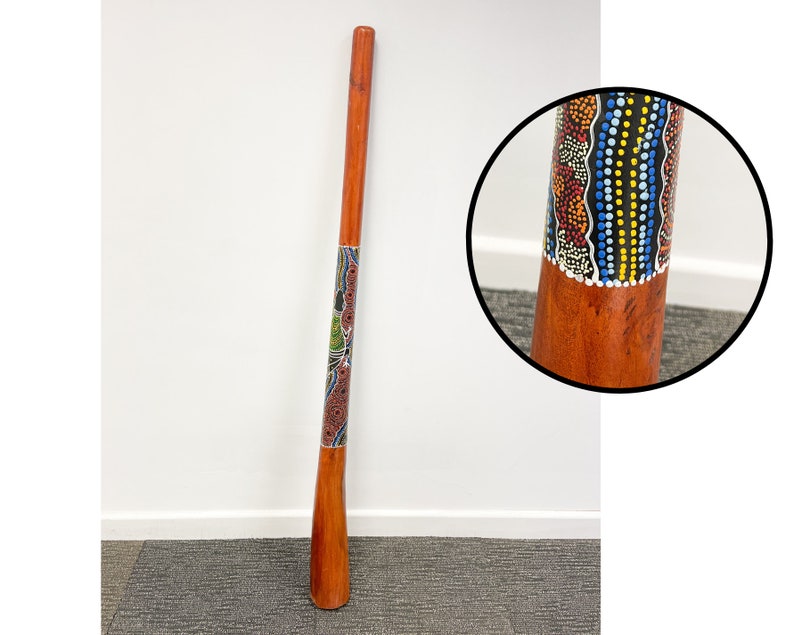 May include: A wooden didgeridoo with a colorful Aboriginal art design. The didgeridoo is painted with red, yellow, blue, black, and white dots and lines. The didgeridoo is about 1 meter long.