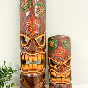 May include: Two wooden tiki masks with carved turtle designs. The larger mask has three turtles, two green and one red, and the smaller mask has two green turtles and one red turtle. Both masks have a brown wood finish and a carved face with a wide mouth and eyes.