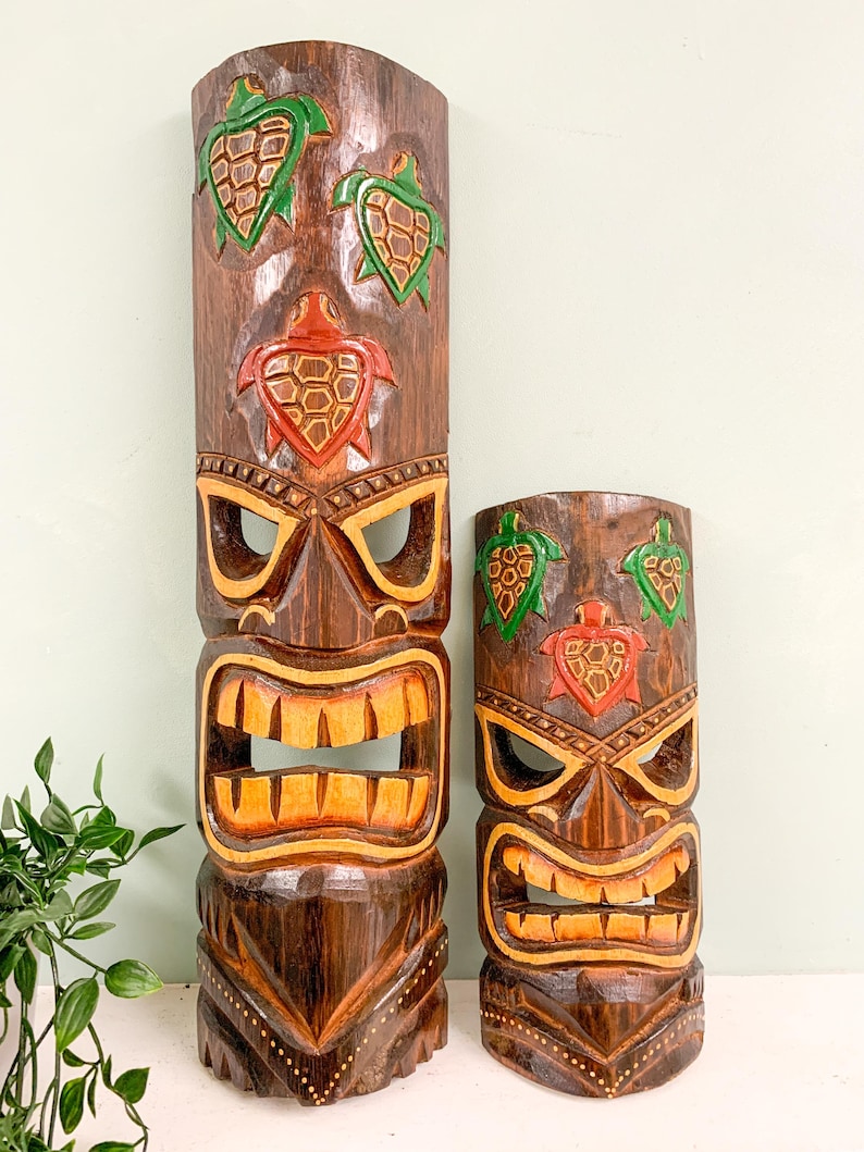 May include: Two wooden tiki masks with carved turtle designs. The larger mask has three turtles, two green and one red, and the smaller mask has two green turtles and one red turtle. Both masks have a brown wood finish and a carved face with a wide mouth and eyes.