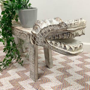 May include: White wooden stool shaped like an alligator head with a carved design. The stool has four legs and is sitting on a white and orange checkered rug.
