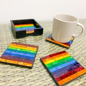 May include: Set of four rainbow-coloured glass coasters with black frames. The coasters are arranged in a square shape with a white mug on top of one of the coasters. The coasters are on a woven wicker surface.