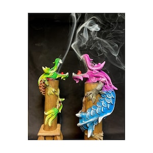May include: Two dragon incense holders, one green and one pink, with blue and white scales, are burning incense. The smoke curls up from the incense holders.