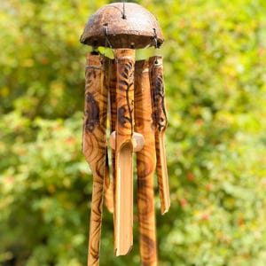 May include: A handmade wind chime with a coconut shell top and bamboo chimes. The bamboo chimes are etched with decorative patterns and hang from a string. The wind chime is a natural brown colour and is suspended against a blurred green background.