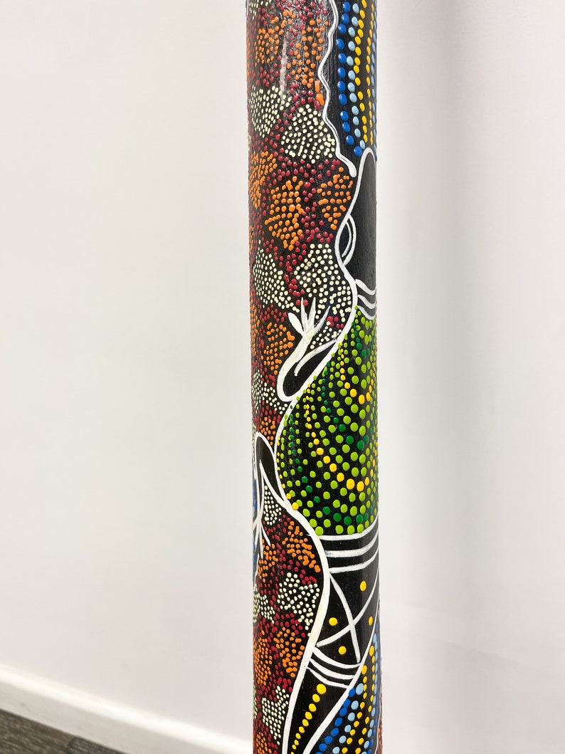 May include: A wooden didgeridoo with a colorful Aboriginal art design. The design features a black, white, green, orange, red, yellow, and blue dot pattern. The didgeridoo is approximately 30 inches long.