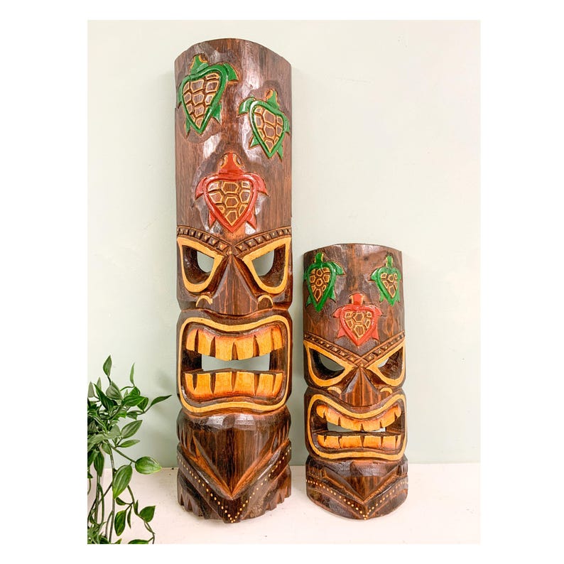 May include: Two wooden tiki masks with carved details and painted accents. The larger mask has a green, red, and brown color scheme, while the smaller mask has a green, red, and brown color scheme. Both masks have a carved turtle design.