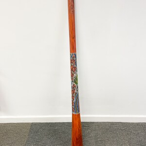 May include: A wooden didgeridoo with a colorful Aboriginal design painted on it. The didgeridoo is about 1.5 meters long and has a natural wood finish.